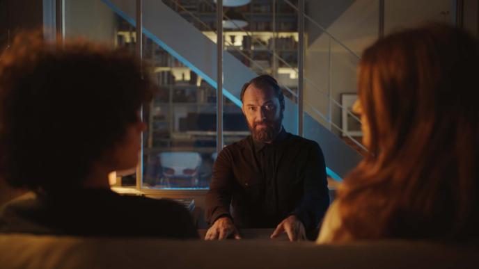 actor jude law looking directly into the camera while sitting in front of two people