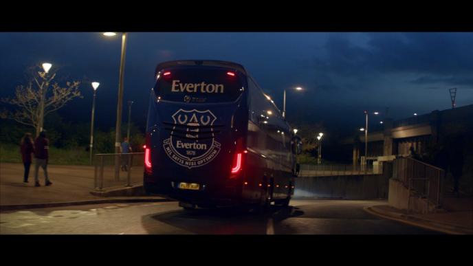 back shot of an everton branded bus driving down a road as three pedestrians walk nearby during nighttime