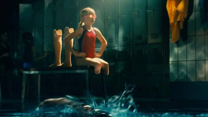a child with an amputation sitting in a power position with their swimsuit and swimming goggles on. their prostheses are behind them. they are in an animated locker room. there are two children in the distance. someone is swimming by.