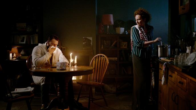 a person sitting at a candle lit table blowing into a tissue. there is another person by the kitchen counter stirring a sauce pan