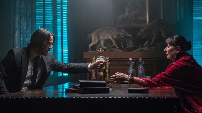 side view of actor keanu reeves as john wick handing actress anjelica huston as the director a ring as they sit opposite each other at a table