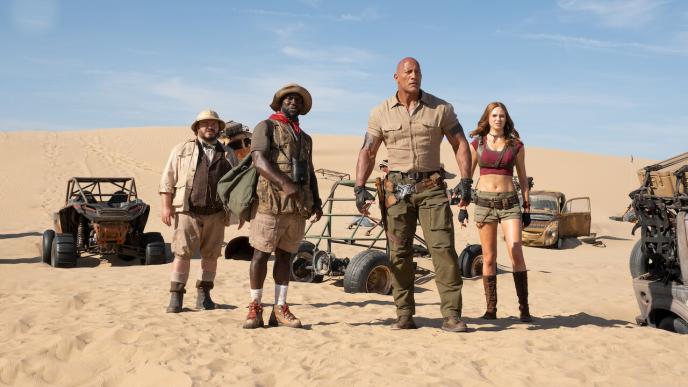 four jumanji characters standing in a desert. there are quad bikes and cars in the background