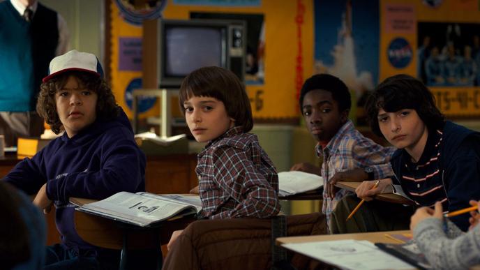 stranger things characters dustin, will, lucas and mike sitting at school desks looking towards the back