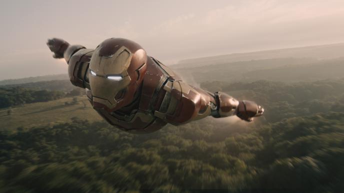 front side view of iron man flying in air with his arms to the sides of his body above forest greenery