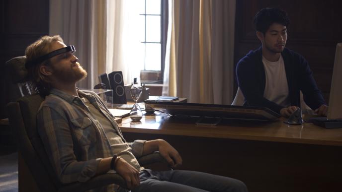 actor wyatt russel as cooper sitting in a chair with a vr headset on as actor ken yamamura as shou sits behind the desk looking at a computer screen