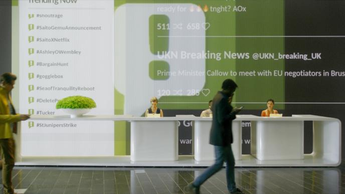 a futuristic reception that has three receptionists in separated tables sitting in front of a wall that covered in a screen projecting social media metrics. two people are walking by
