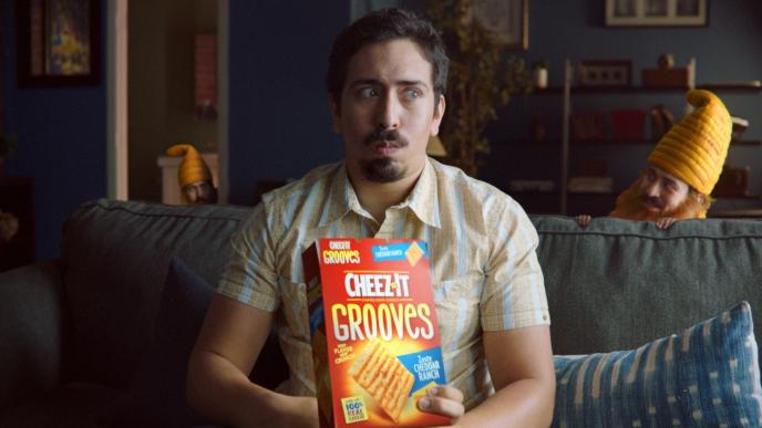 a person sitting on a sofa eating cheez-it grooves crackers as two cheese gnomes stand behind