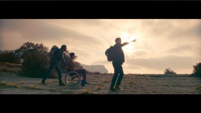 three people hiking through mountainous terrain. one of them is pointing ahead, one is in a wheelchair and the other one is supporting the person in the wheelchair