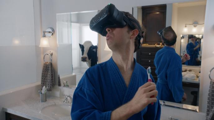 a man wearing a vr headset holding a toothbrush in a bathroom as a cow peaks its head through the door