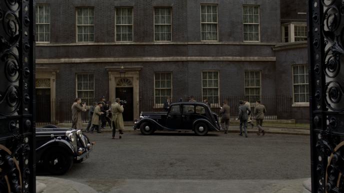 set in the 1940s, journalists in trench coats taking photos of a car as men in suits rush towards the car door