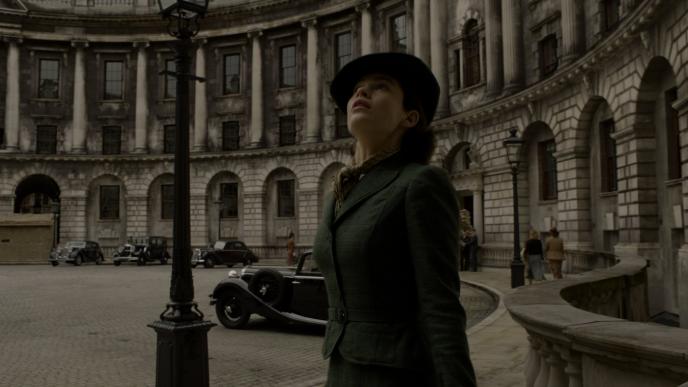 actress lily james as elizabeth layton weating a dark green dress suit and hat looking up in front of a war torn dirty downing street