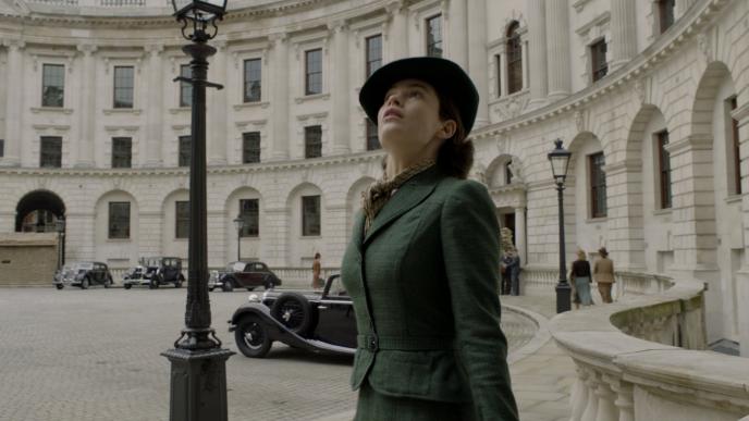 actress lily james as elizabeth layton weating a dark green dress suit and hat looking up in front of downing street