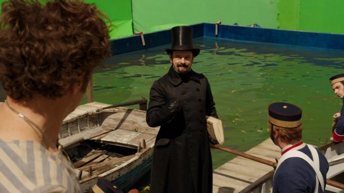 actor michael sheen as dr blair mudfly standing in front of a green screen as actors stand next to him