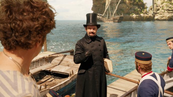 actor michael sheen as dr blair mudfly standing in front of sea as actors stand next to him