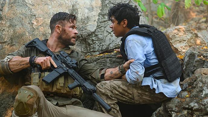 actors chris hemsworth and rudhraksh jaiswal crouched in front of a boulder. chris hemsworth is holding a gun as he is holding onto rudhraksh jaiswals arm