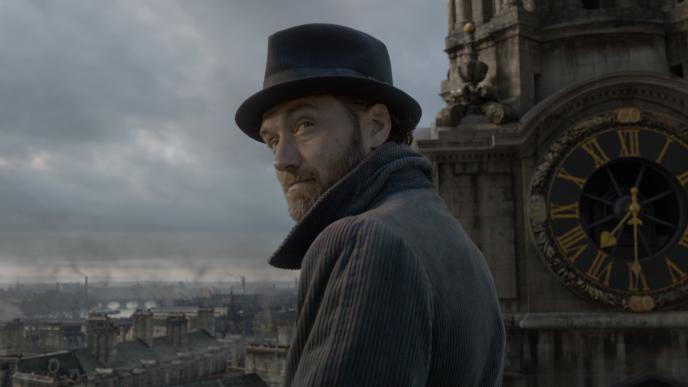 actor jude law as albus dumbledore looking back. he is wearing a fedora and grey jacket. he is standing next to a clock tower