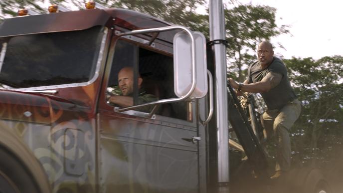 actor jason statham as deckard shaw driving a lorry and dwayne johnson as luke hobbs holding on the back of the cab side