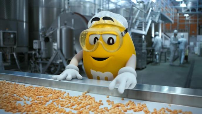 cg animated yellow m&m pointing at a conveyor belt full of peanuts