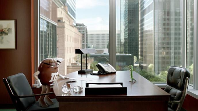 cg animated geico gecko and chocolate m&m having a discussion at an office desk as the window overlooks cityscapes