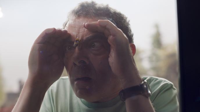 actor rowan atkinson looking through a glass window with a bee perched in the middle of his eyebrows