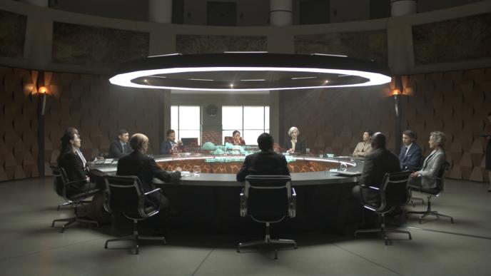 a circular table with a hologram projector circling the top of the table. there are twelve people in suits sitting at the table looking at a hologram in the centre of the table