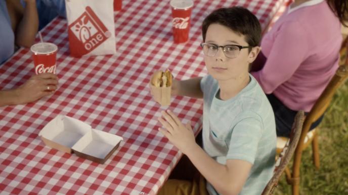 a child sitting at a picnic table, looking up to the camera while holding a chick-fil-a burger