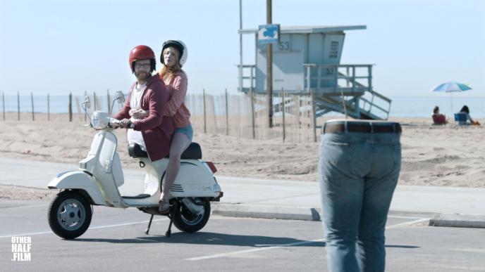 a man's torso at the front of a vespa motorbike and a woman sitting on the back of the bike. the man's legs are standing facing the man to the right