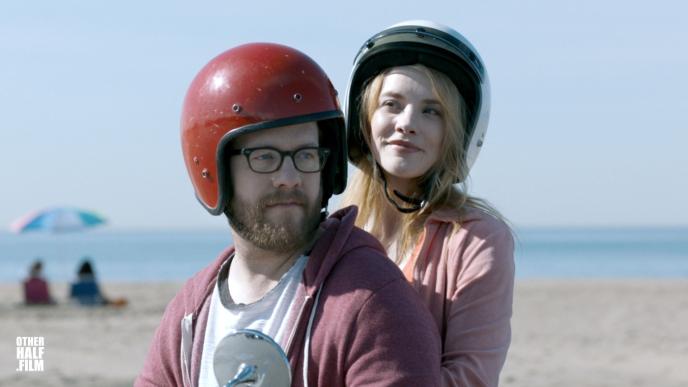 close up of a man and a woman wearing helmets. there is a beach and sea in the background