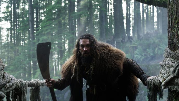 actor jason momoa as character baba voss wearing a fur jacket holding a machete peering into a cave