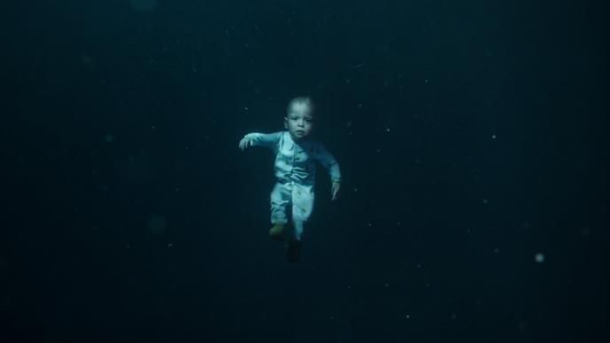 a baby floating underwater in deep dark waters looking directly into the camera