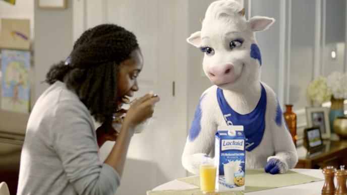 a woman eating cereal as a cg animated cow character smiles at her