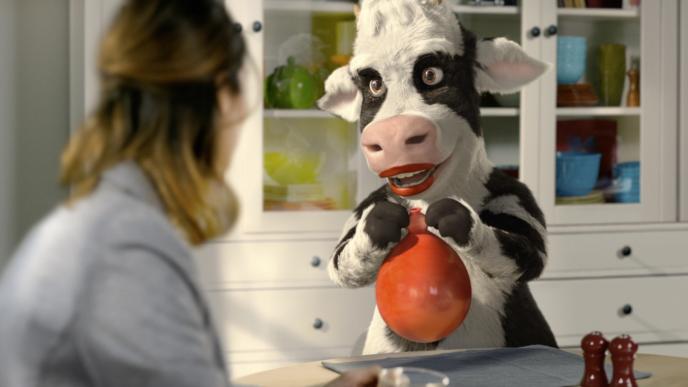 a cg animated cow character holding a balloon full of milk as a woman drinks coffee