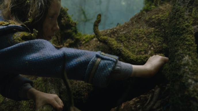 actress dixie egerickx as mary lennox holding a mossy tree branch and looking at a mossy tree stump