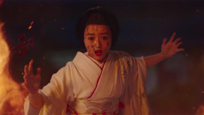 a geisha wearing white robes lined with red linen staring with a look of terror on her face as kanji characters appear on her face. there are flames arising to her left