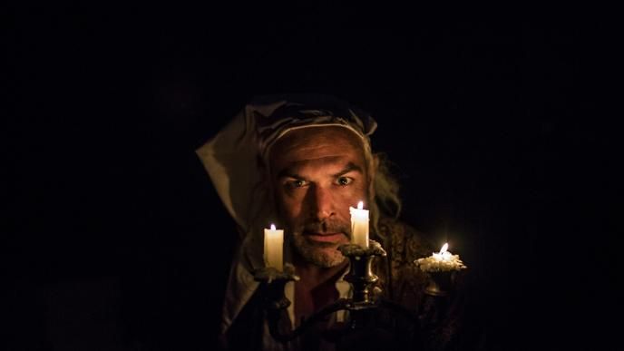 a victorian man wearing night pyjamas holding a candle holder with three lit candles in darkness