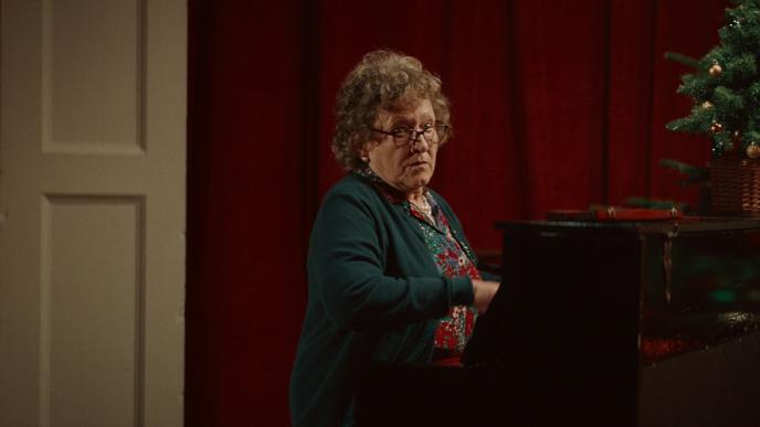 a grandma sitting at a piano looking over towards the camera. there is a christmas tree on the right side of the piano