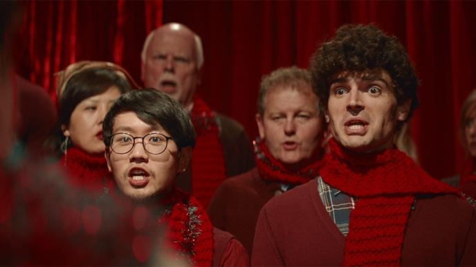 a christmas choir wearing red scarves and jumpers singing hastily while looking confused