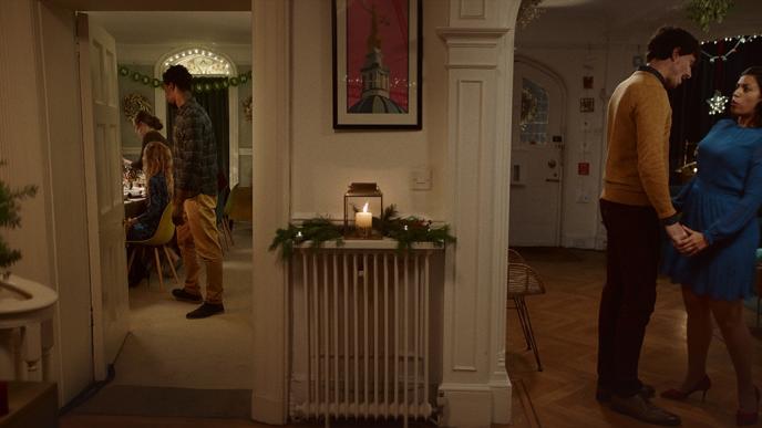 a couple standing under mistletoe in a hallway as people visible through a door are gathered in the distance on the left in a kitchen
