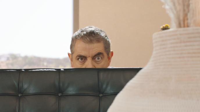 actor rowan atkinson looking ominously from behind a sofa