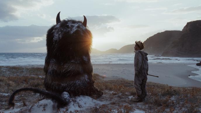 back view of cg animated carol the monster sitting down and max standing next to the monster in front of the sea at a beach at sunset