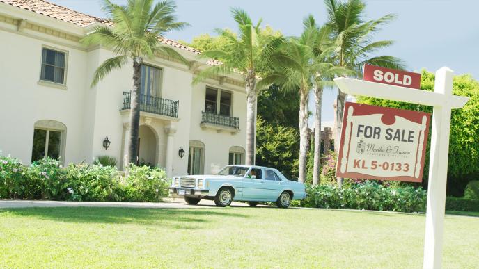 a pasadena mansion in a gated community that has palm trees at the front. there is a blue mustand car parked by the driveway and there is a board up that indicates the mansion is up for sale