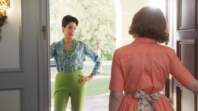 back view of a woman wearing a coral dress with a floral bow tie opening the door as another woman stands with her hand on her hip wearing a purple and green blouse, green bead necklace and lime green trousers