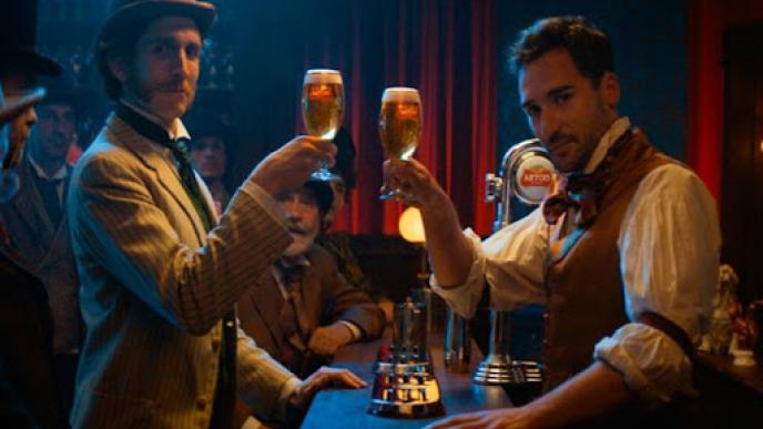 two actors wearing victorian clothing holding up pints of beer while looking into the camera amongst a crowd in a bar