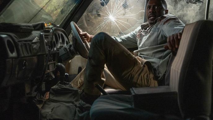 Idris Elba braces against a car door with a smashed window, gripping the steering wheel