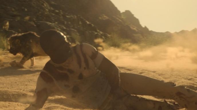 Idris Elba lies on the ground as a lion stalks in the background