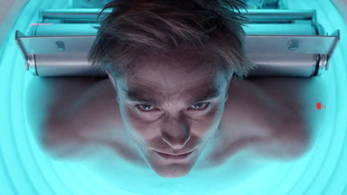 A blonde man lies in a machine, illuminated with light blue lights