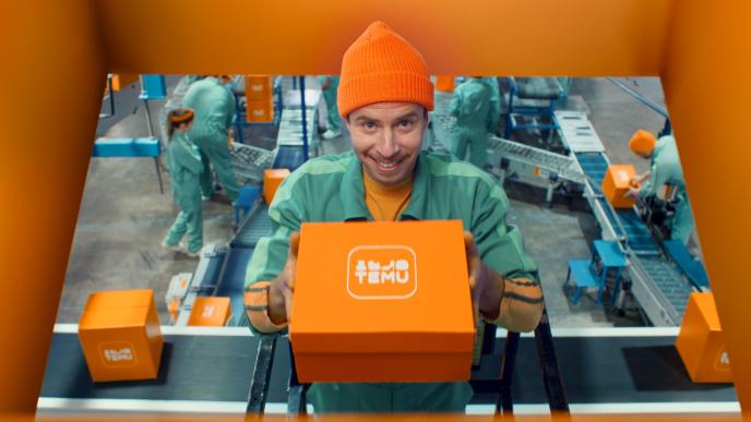 A man in an orange beanie hat and turquoise jacket holds an orange box