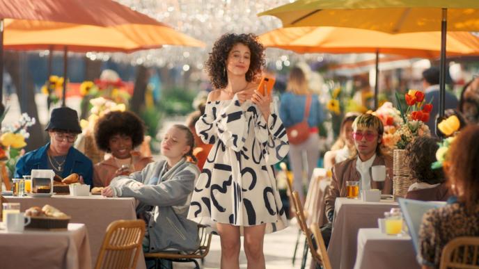 A young woman in a black and white dress walks through an outdoor seating area with yellow parasols