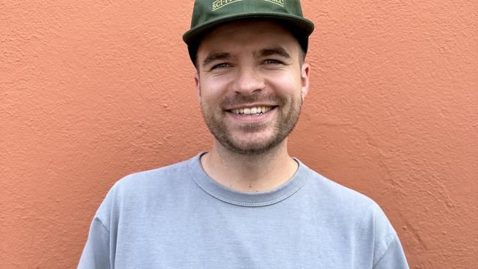 A headshot of Senior Producer, James Yeo. He is wearing a green cap and a grey sweatshirt, stood against an orange wall.