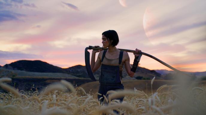 A young woman stands in a field, the sun is setting, she balances a scythe over her shoulders